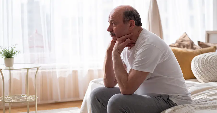 Trauma Solutions: Man with gray hair and beard sits on a bed, leaning forward with his hands clasped near his mouth, appearing deep in thought.