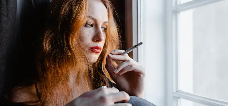 Trauma Solutions: A woman with long red hair sits by a window, holding a cigarette and looking outside with a contemplative expression, lost in mindful reflection and thoughts of self-esteem. A woman sits holding a lit cigarette while staring out of the window. Many compulsive and addictive behaviors like smoking find their roots in attachment disruptions, as people seek substitute connection with substances and other behaviors.
