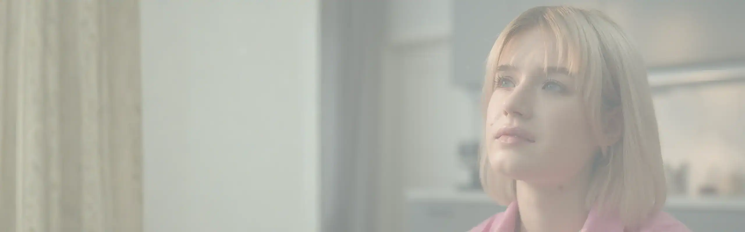 Trauma Solutions: A woman with short blonde hair and a pink shirt gazes upward, sitting indoors in a moment of mindfulness, with a blurred background of curtains and kitchen cabinets. Thoughtful young woman representing the emotional experience of addiction, attachment wounds, and the search for connection.