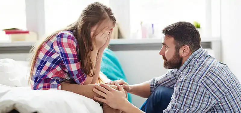 Trauma Solutions: A man kneels beside a bed, consoling a young woman who is sitting with her face in her hands, appearing upset.