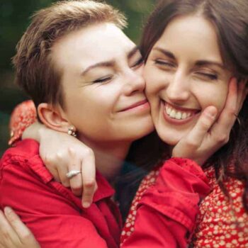 Trauma Solutions: Two women in red clothing smile as one hugs and gently holds the other’s face outdoors, both appearing happy and affectionate.