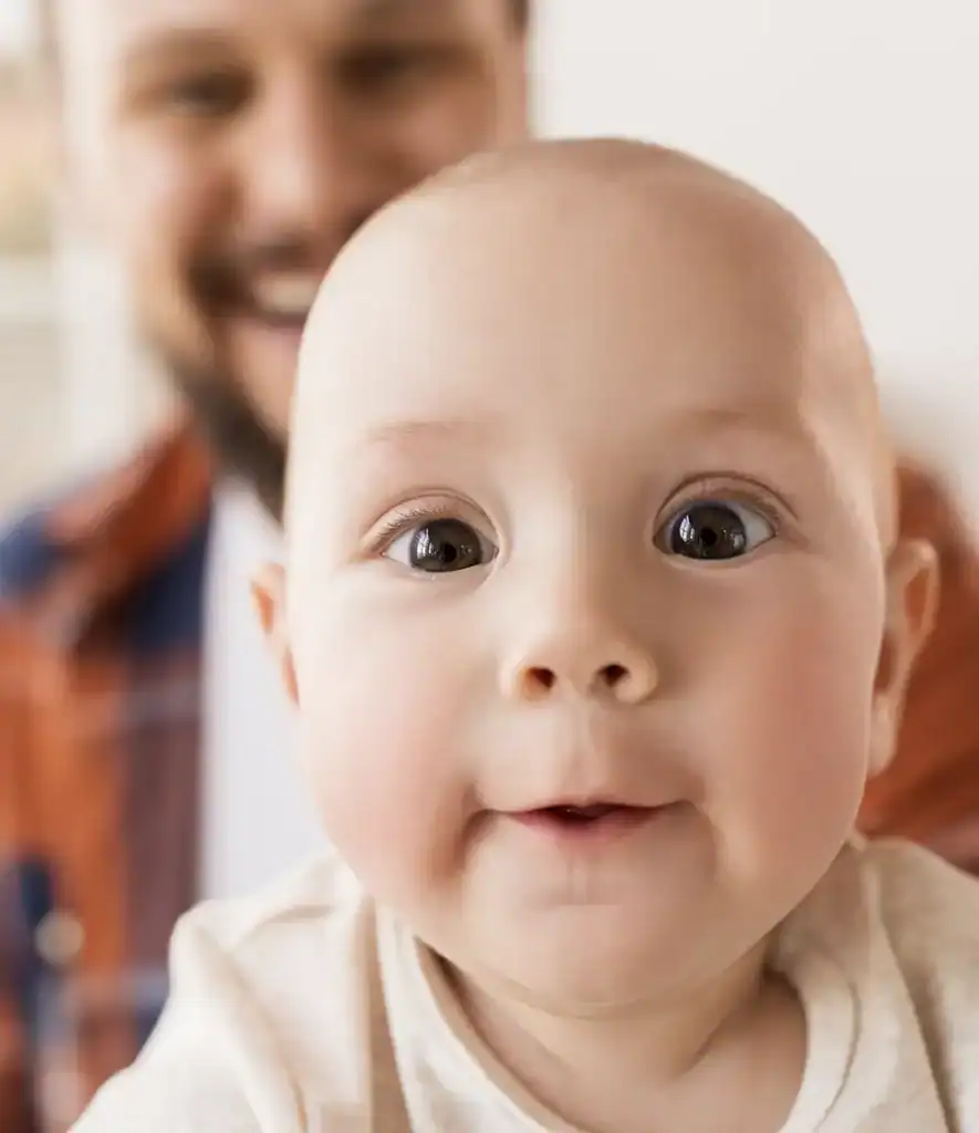 Trauma Solutions: A close-up of a baby with wide eyes and a slight smile, with an adult male figure smiling in the blurred background.