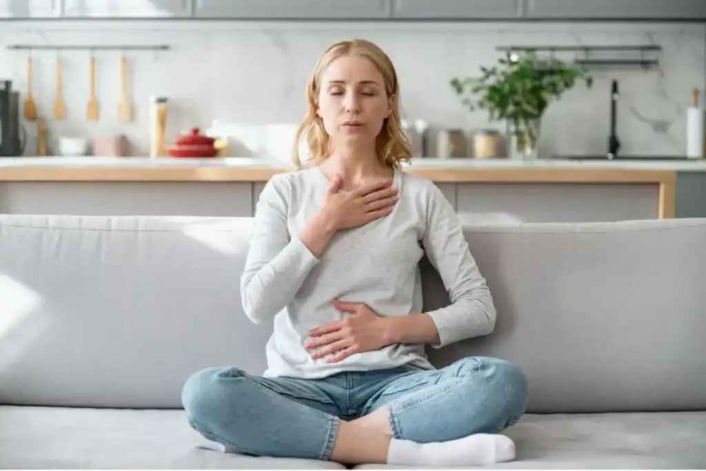 Trauma Solutions: Woman sits cross-legged on a couch, eyes closed, with one hand on her chest and the other on her abdomen, appearing to practice mindfulness or meditation to boost self-esteem.
