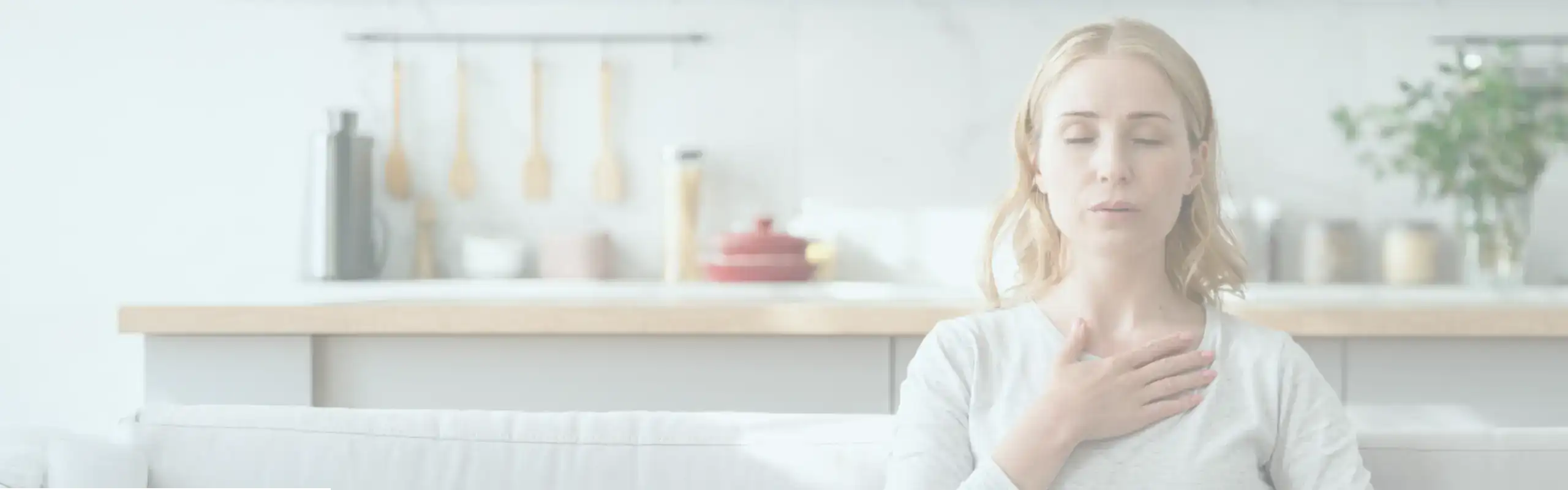 Trauma Solutions: A woman sits on a couch with her eyes closed and one hand on her chest, practicing mindful deep breathing in a bright kitchen setting.