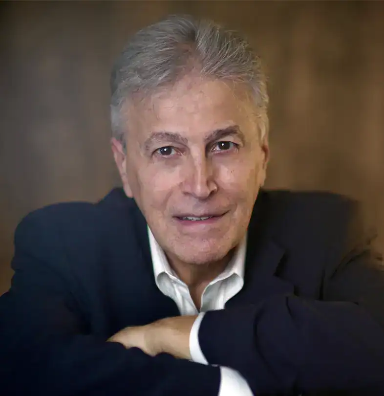 Trauma Solutions: Older man with gray hair in a dark suit jacket and white shirt, sitting with arms crossed on a table, looking at the camera with a neutral expression against a blurred brown background.
