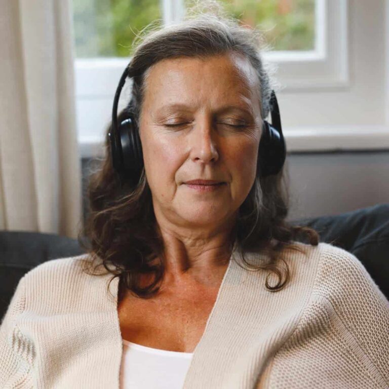 Trauma Solutions: Middle-aged woman with closed eyes sits on a couch, wearing black headphones and a beige sweater, appearing relaxed.