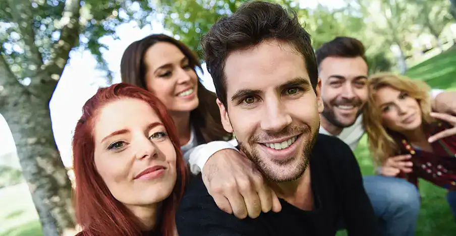 Trauma Solutions: Five people sit closely together outdoors on grass, smiling at the camera. Trees and greenery are visible in the background.