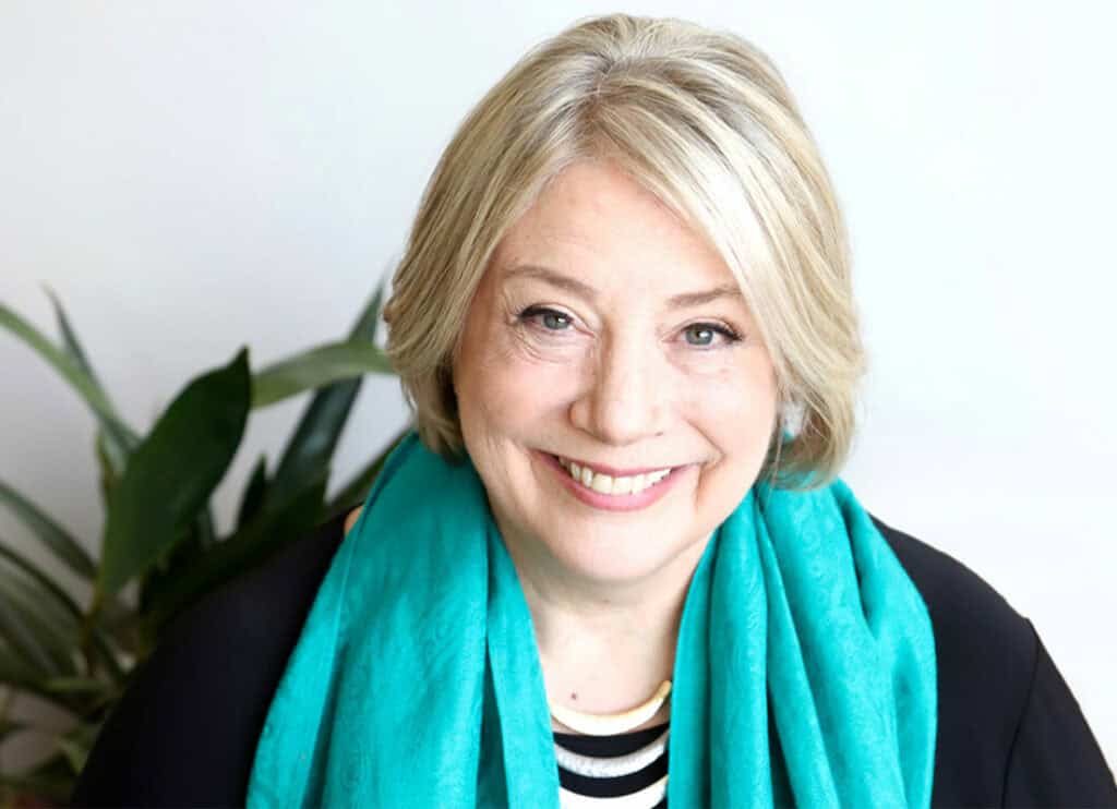 Trauma Solutions: Smiling older woman with short blonde hair wearing a turquoise scarf and black top, sitting in front of a white background with a plant visible on the left.