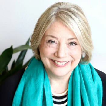 Trauma Solutions: Smiling older woman with short blonde hair wearing a turquoise scarf and black top, sitting in front of a white background with a plant visible on the left.