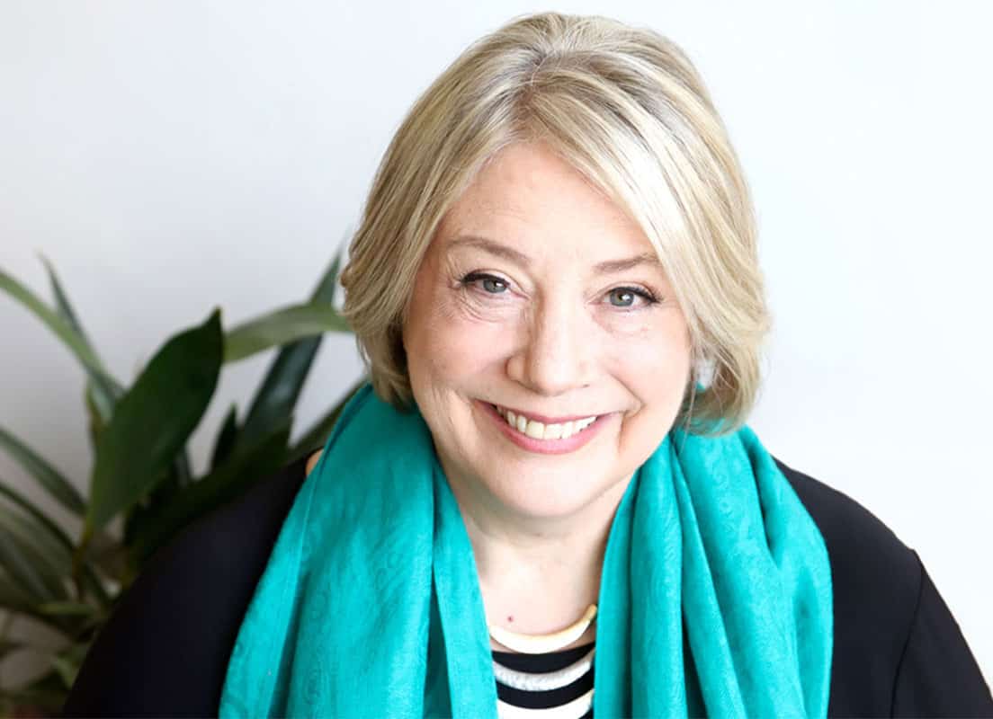 Trauma Solutions: Smiling older woman with short blonde hair wearing a turquoise scarf and black top, sitting in front of a white background with a plant visible on the left.