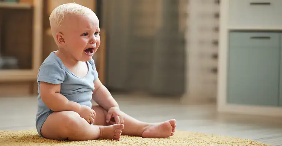Trauma Solutions: A baby with blonde hair sits on a yellow rug indoors, crying with an open mouth and tears on their face.