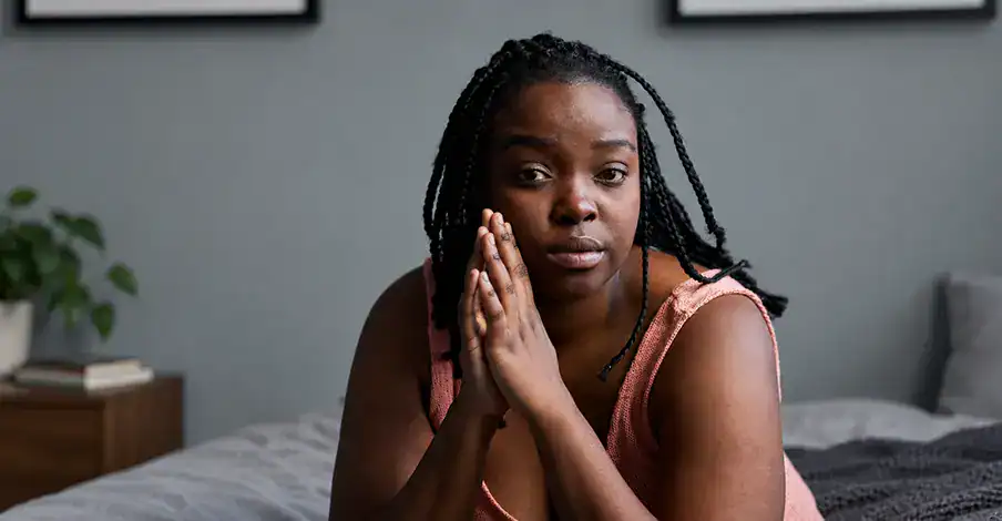 Trauma Solutions: A woman with braided hair sits on a bed, leaning forward with her hands together, looking directly at the camera in a softly lit room.