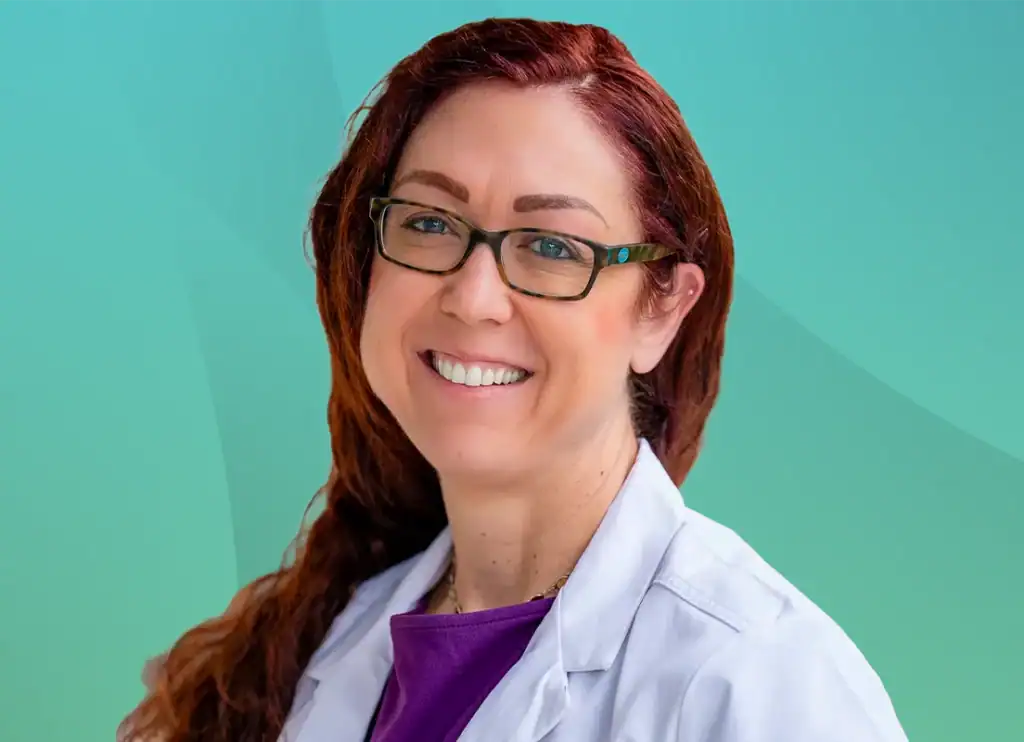 Trauma Solutions: A person with long red hair and glasses smiles while wearing a white lab coat in front of a teal and green background.
