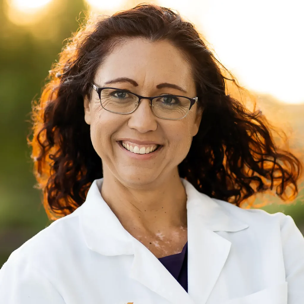 Trauma Solutions: Woman with wavy brown hair wearing glasses and a white lab coat smiles outdoors with a blurred green background.