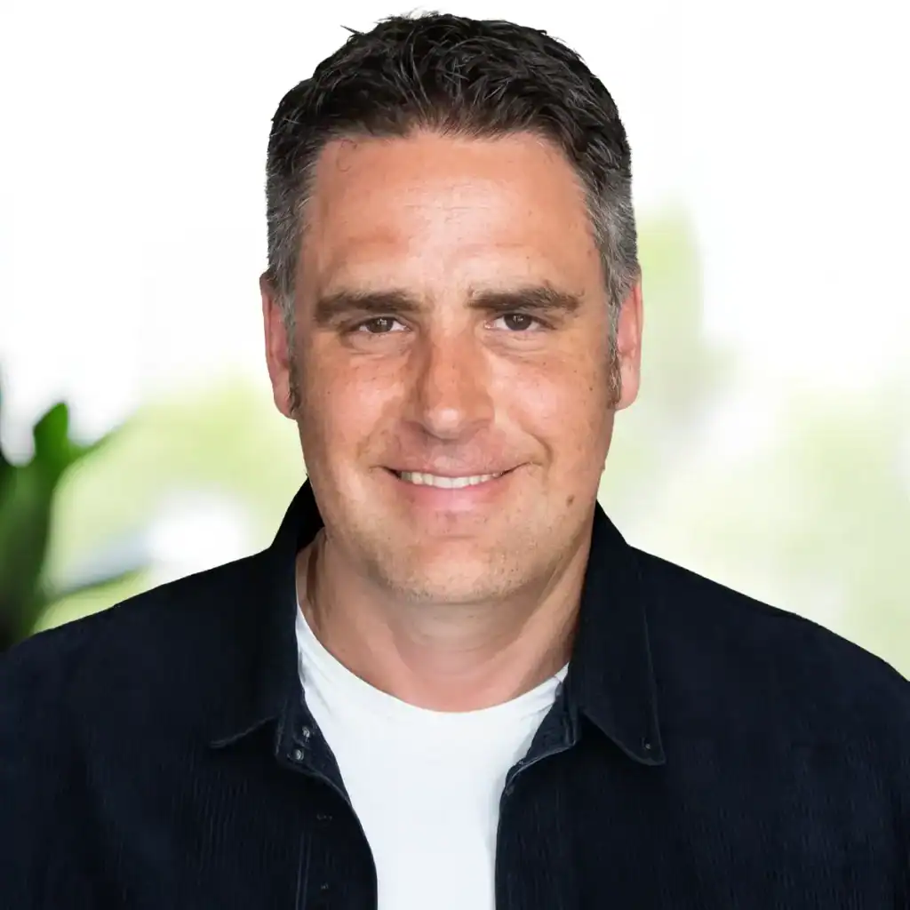 Trauma Solutions: A man with short dark hair wearing a white t-shirt and dark jacket smiles at the camera in a brightly lit indoor setting.