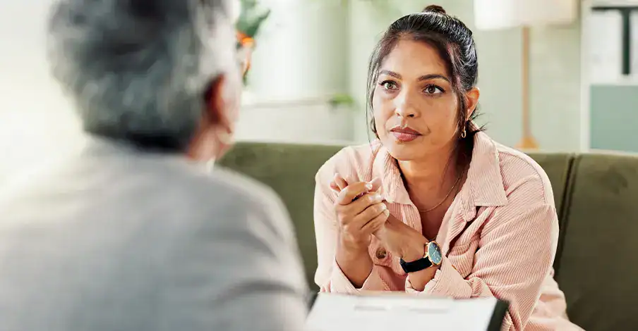 Trauma Solutions: A woman sits on a couch, listening attentively to another person whose back is to the camera.