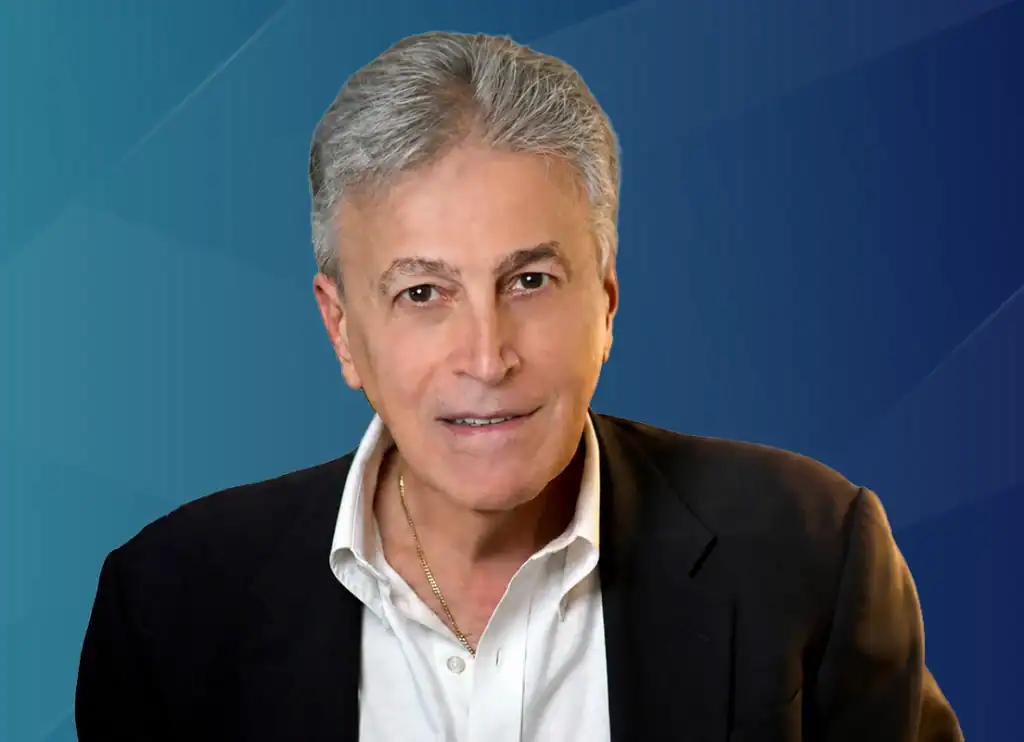 Trauma Solutions: A man with gray hair wearing a dark blazer, white collared shirt, and gold necklace, poses in front of a blue geometric background.