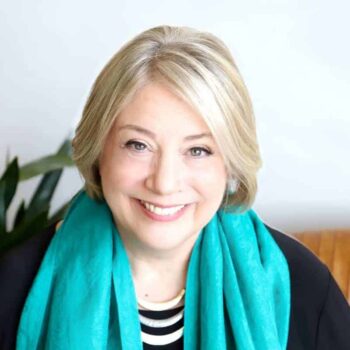Trauma Solutions: A woman with short blonde hair smiles at the camera, wearing a turquoise scarf over a black and white striped top, with a plant in the background.
