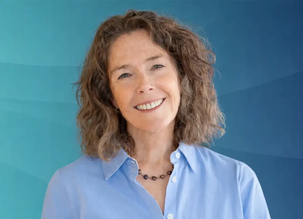 Trauma Solutions: Dr. Liz George with curly brown hair, wearing a light blue button-up shirt and a black beaded necklace, smiles in front of a blue gradient background.