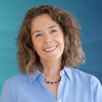Trauma Solutions: Dr. Liz George with curly brown hair, wearing a light blue button-up shirt and a black beaded necklace, smiles in front of a blue gradient background.