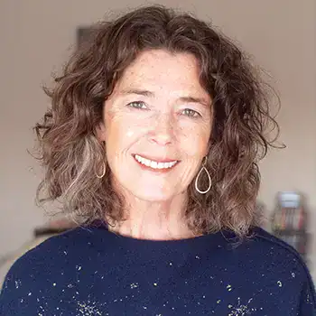 Trauma Solutions: Smiling woman with curly brown hair wearing drop earrings and a dark sweater, standing indoors with a blurred background.