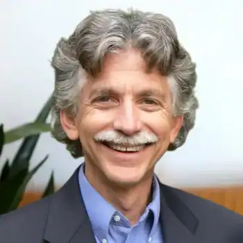 Trauma Solutions: A middle-aged man with curly gray hair and a mustache, wearing a blue shirt and dark blazer, smiling in front of a blurred background with a plant.