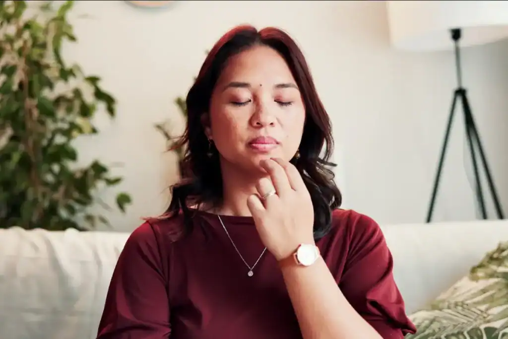 Trauma Solutions: A woman with closed eyes sits on a couch touching her chin with her fingers, appearing calm and embodying mindfulness. Indoor setting with plants and a lamp in the background.