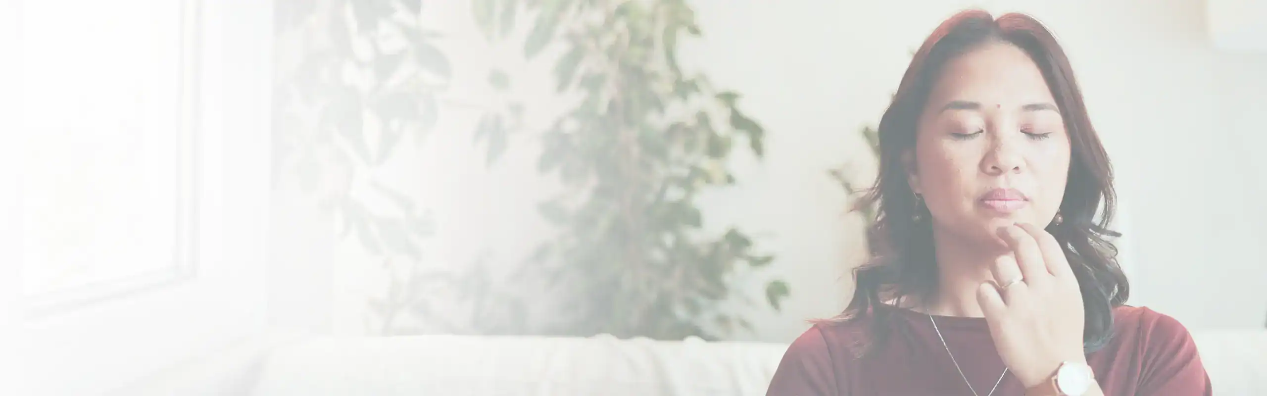 Trauma Solutions: Woman with closed eyes touches her chin with two fingers, sitting indoors near a plant, appearing calm and focused, embodying mindfulness and self-esteem.