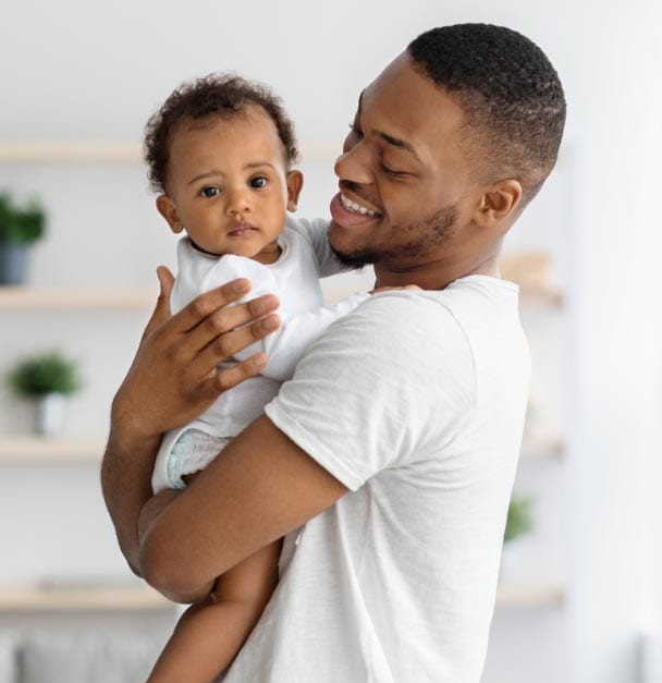 A black man practicing the DARe (Diversity and Racial Equality) approach, holding a baby in his arms in a living room.