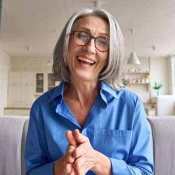 Trauma Solutions: Smiling older woman with gray hair and glasses wearing a blue shirt, sitting indoors in a bright, modern kitchen.