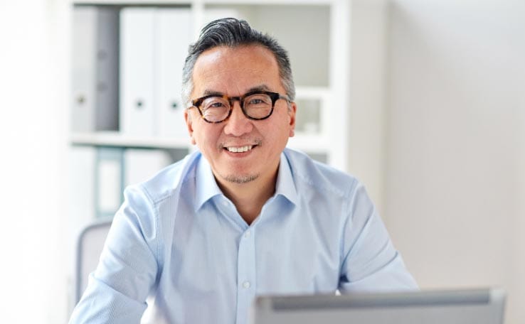 A smiling Asian man mastering trauma at a desk with a laptop.