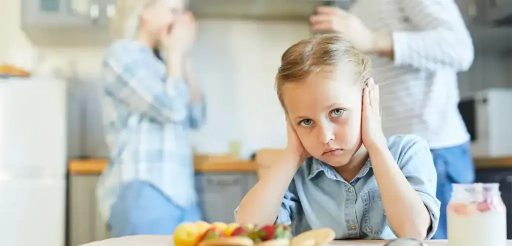 Trauma Solutions: A young girl sits at a kitchen table covering her ears, looking unhappy, while two adults talk in the background.