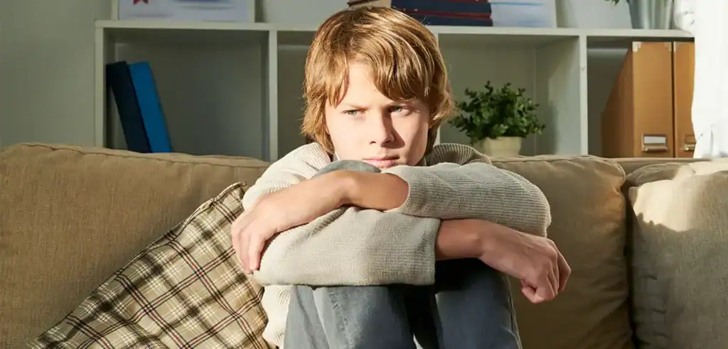 Trauma Solutions: A young person with light brown hair sits on a couch, hugging their knees and looking thoughtfully into the distance.