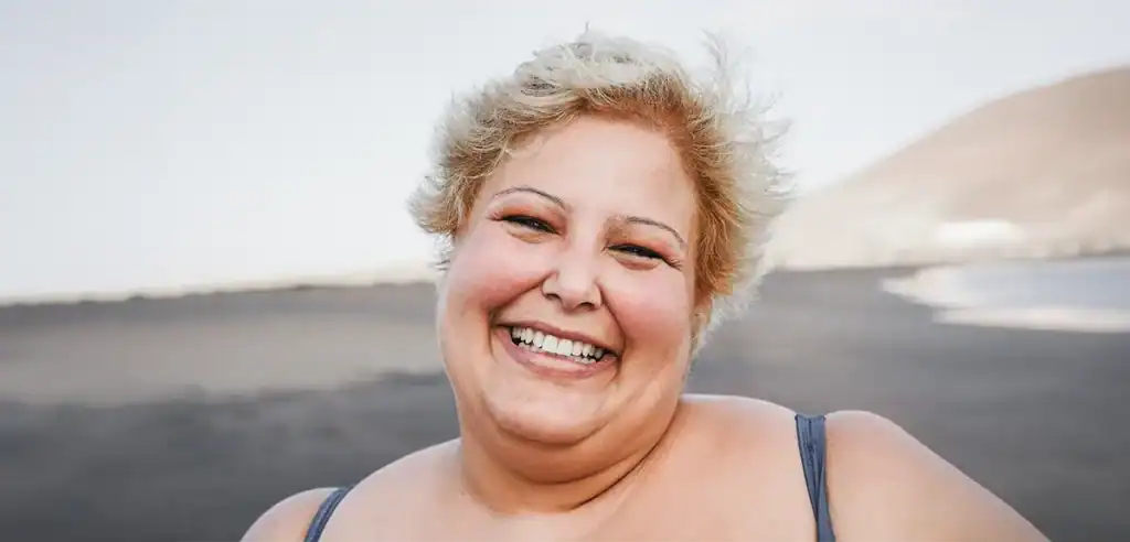 Trauma Solutions: A person with short blonde hair smiles outdoors on a beach, with sand and water visible in the background.