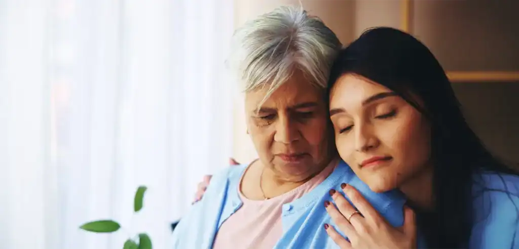Trauma Solutions: An older woman and a younger woman sit close together. The younger woman rests her head on the older woman's shoulder with her eyes closed, both wearing light blue clothing.