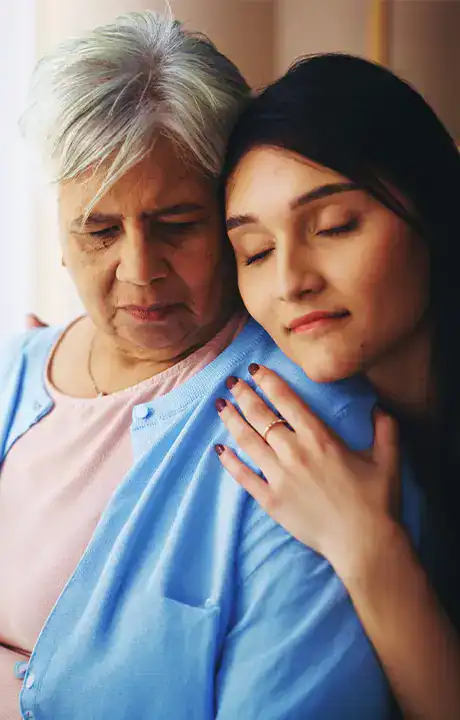 Trauma Solutions: An older woman and a younger woman sit closely together. The younger woman rests her head on the older woman's shoulder with her eyes closed, both wearing calm expressions.