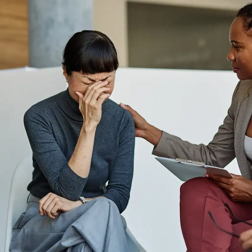 Trauma Solutions: A woman sits with her hand covering her face while another person with a clipboard gently touches her shoulder in support.