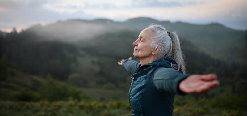 A woman with silver hair stands on a hillside at dusk, arms outstretched and eyes closed in a peaceful expression. Her posture embodies the kind of somatic release and embodied freedom that can emerge through healing attachment patterns, showing both physical expansion and emotional openness.