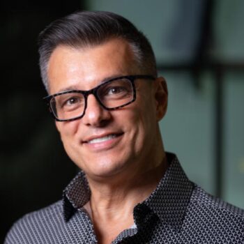 Trauma Solutions: A man with short gray hair, wearing black-rimmed glasses and a patterned shirt, smiles at the camera with a blurred background.