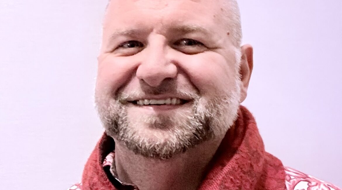 Trauma Solutions: A man with short blond hair and a beard, wearing a red patterned shirt and a red scarf, smiling in front of a plain light background.