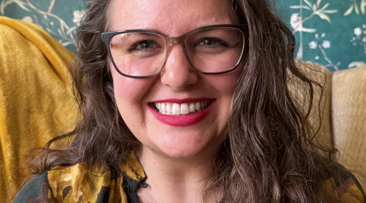 Trauma Solutions: A woman with wavy brown hair, glasses, and a yellow floral top smiles in front of a green botanical wallpaper.