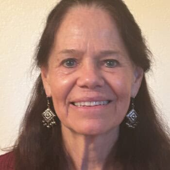 Trauma Solutions: A woman with long brown hair, wearing a red top and dangling earrings, smiles at the camera against a plain light-colored background.