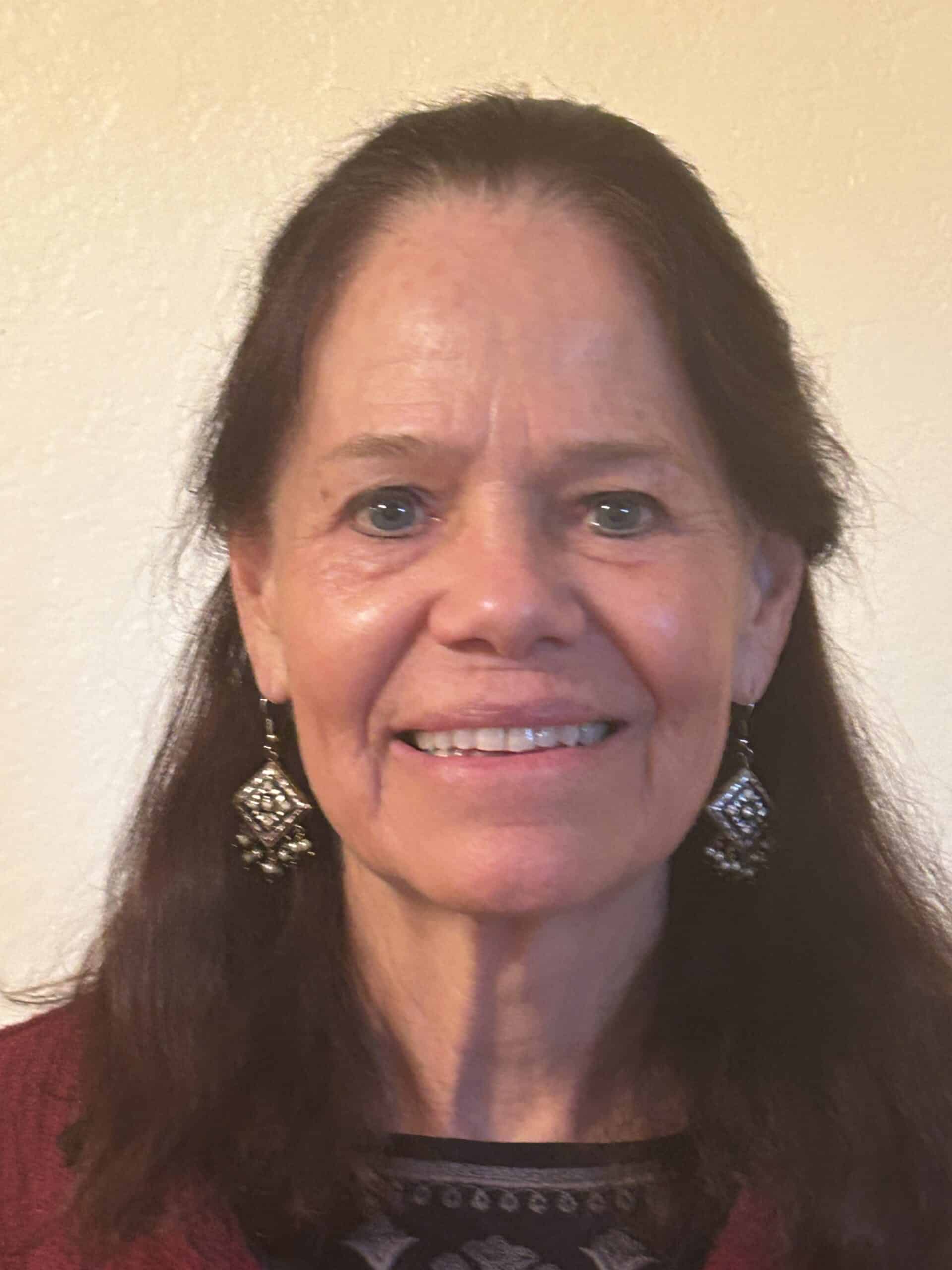 Trauma Solutions: A woman with long brown hair, wearing a red top and dangling earrings, smiles at the camera against a plain light-colored background.