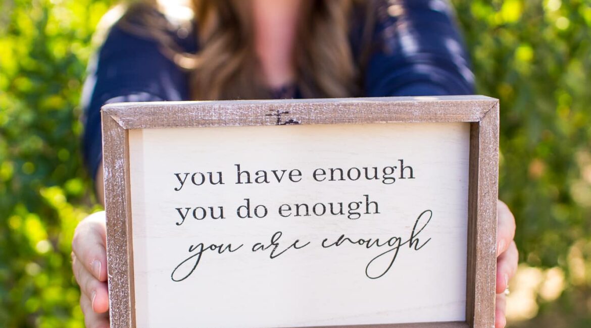 Trauma Solutions: A woman holds a framed sign that reads, “you have enough you do enough you are enough,” with a blurred outdoor background.
