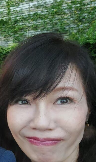 Trauma Solutions: A woman with long dark hair and light makeup is smiling slightly while looking at the camera, with green foliage in the background.