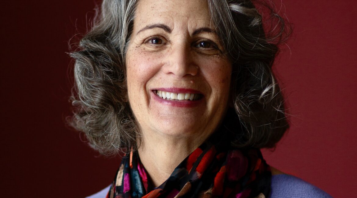 Trauma Solutions: A woman with gray, curly hair and a multicolored scarf smiles at the camera against a dark red background.