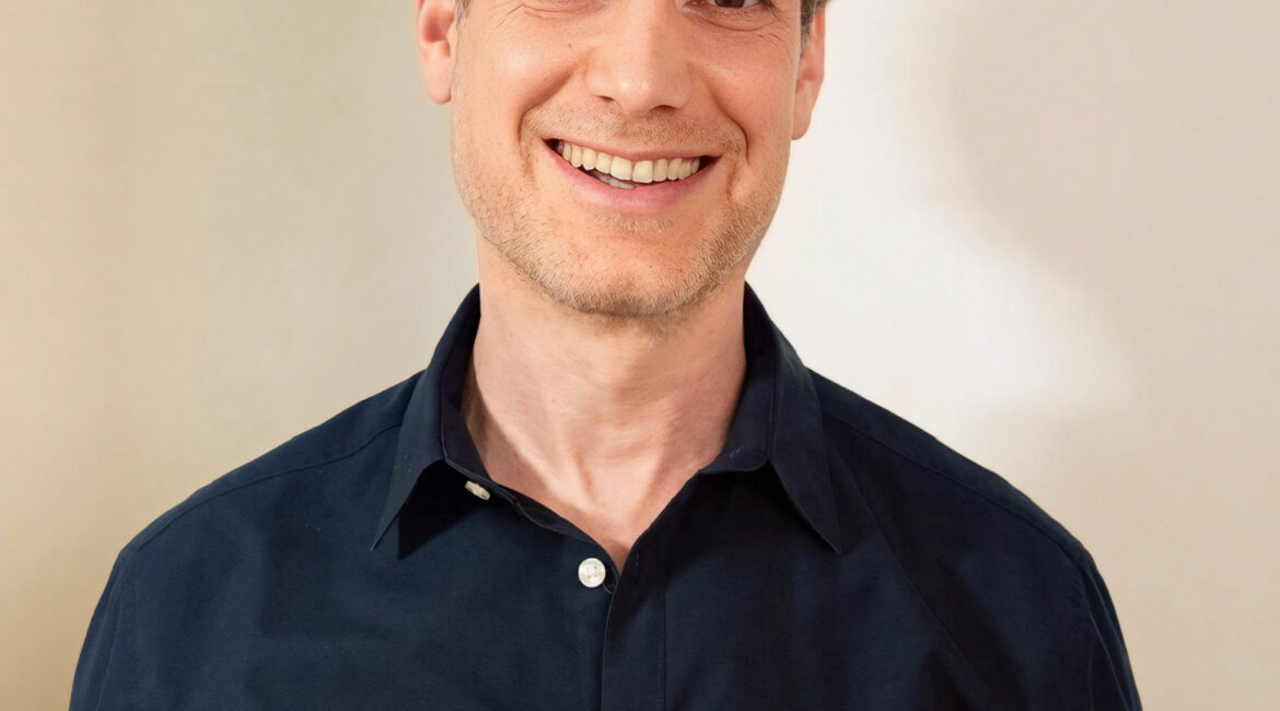Trauma Solutions: A man with short curly hair wearing a dark blue button-up shirt stands in front of a plain, light-colored background, smiling at the camera.