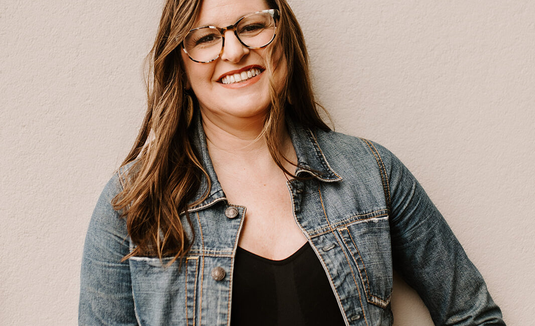 Trauma Solutions: Woman with long brown hair wearing glasses, a denim jacket, and maroon pants stands against a plain light-colored wall, smiling at the camera.