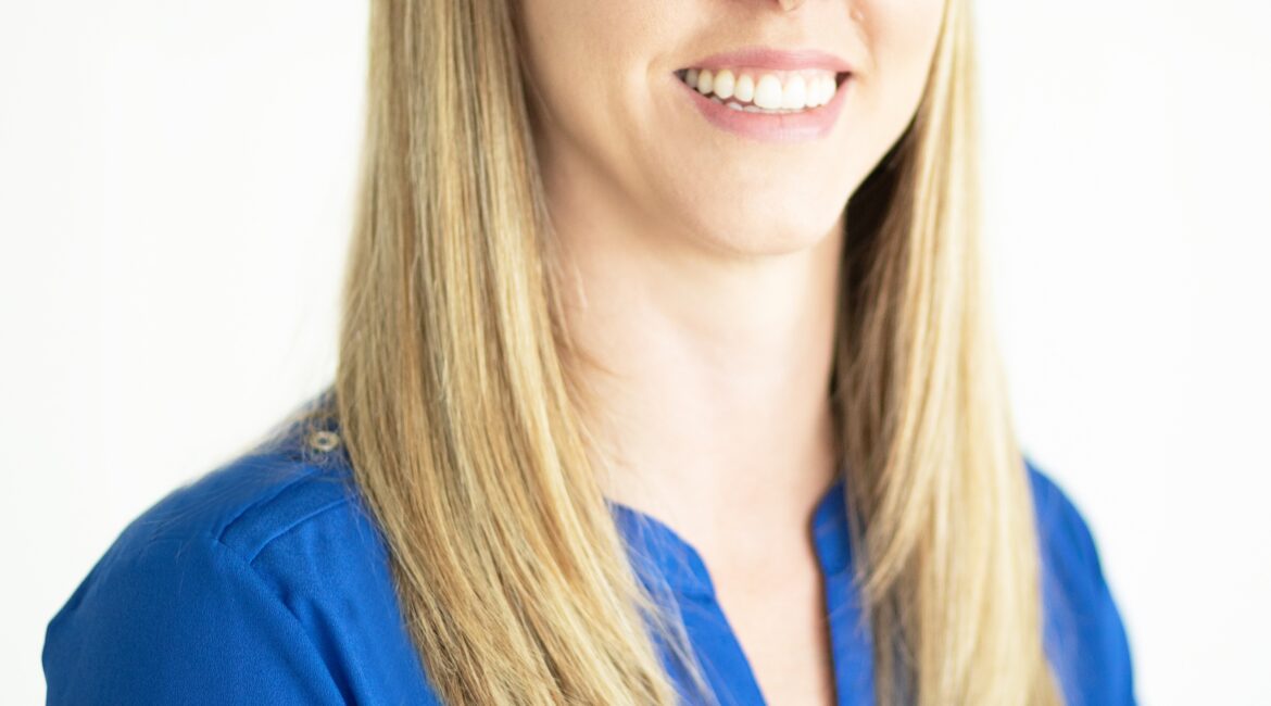 Trauma Solutions: Een vrouw met steil blond haar en een felblauwe blouse staat voor een effen witte achtergrond en lacht naar de camera.