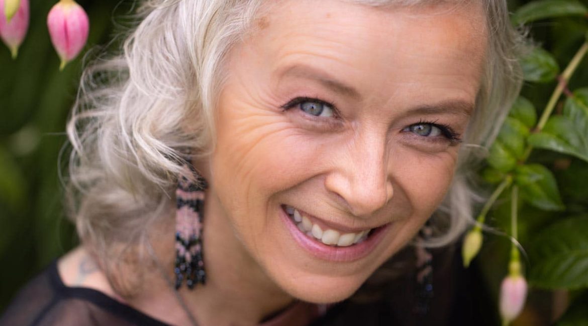A woman with gray hair smiling in front of flowers.